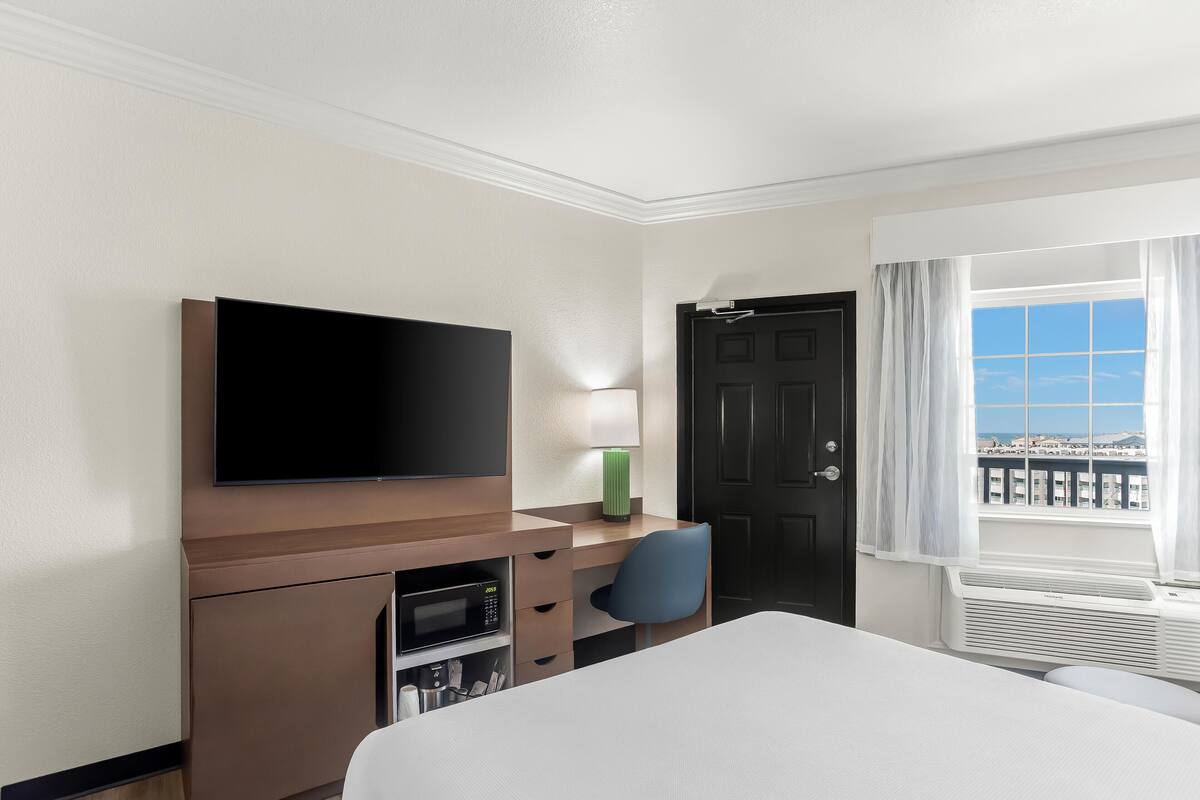 Quarto de hotel com cama, uma escrivaninha com televisão de tela plana, uma cadeira, abajur, porta e janela com vista.