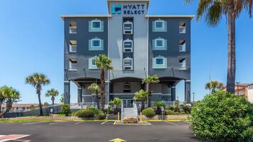 A multi-story hotel with a sign reading 'Hyatt Select,' featuring a central entrance with steps, surrounded by palm trees and a parking lot.