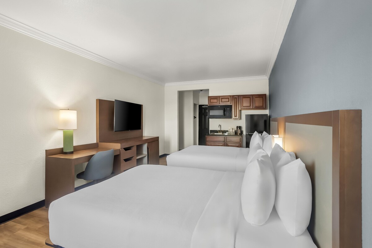 A hotel room with two beds, a desk with a chair, a TV, and a kitchenette.