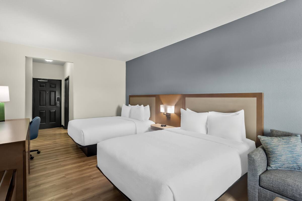 Quarto de hotel com duas camas, uma escrivaninha, uma cadeira e uma mesa de cabeceira com abajur.