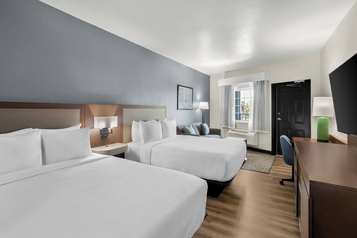 Quarto de hotel com duas camas, uma escrivaninha, uma cadeira, um abajur e uma porta.