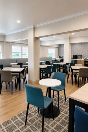 A modern dining area with round tables and chairs, a kitchen in the background, and large windows. - Hyatt Select Galveston - Beachfront (Galveston)