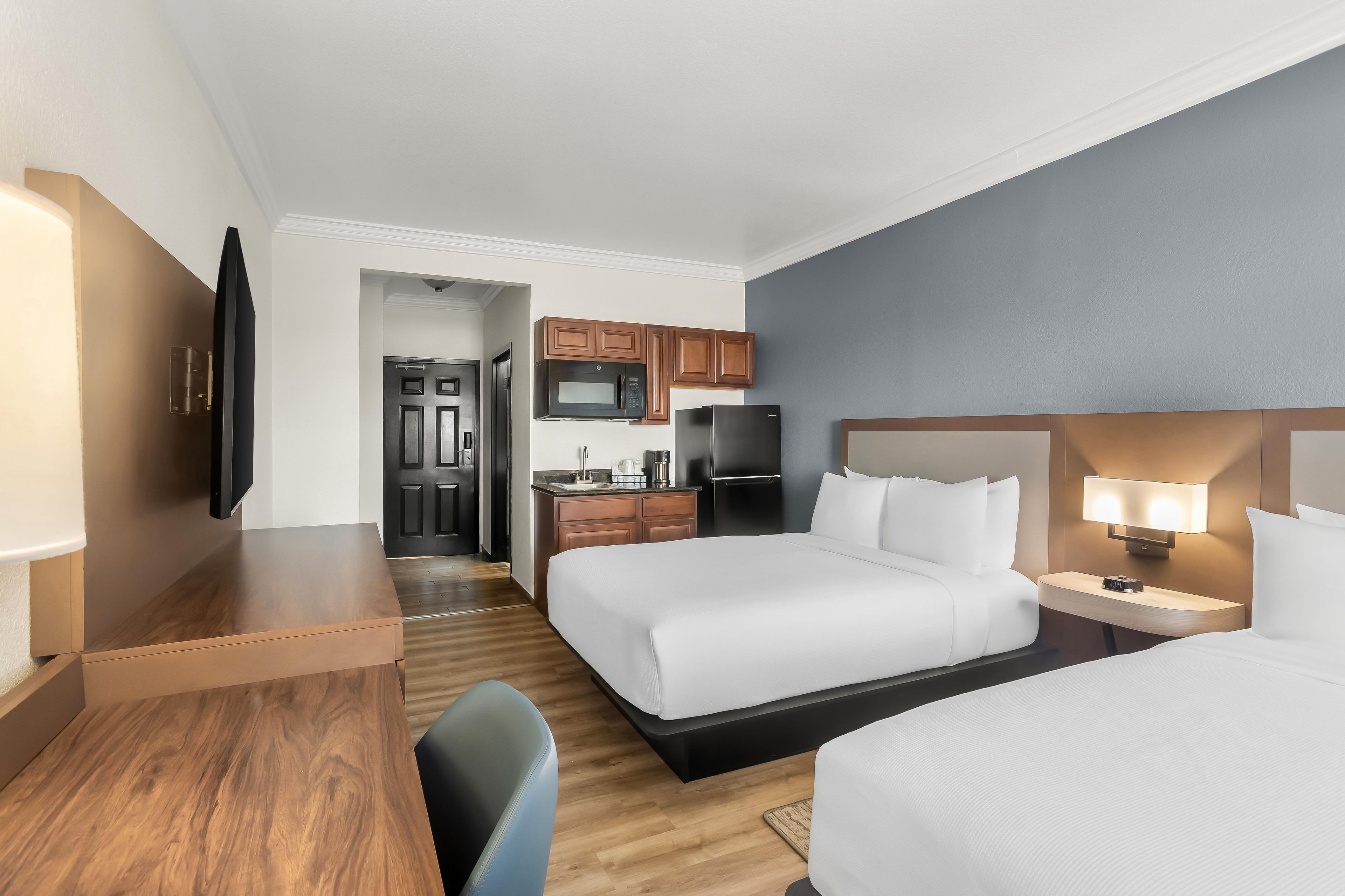 A hotel room with two beds, a desk, a TV, a kitchenette, and a bathroom.