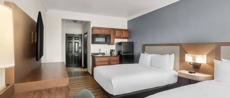 A hotel room with two beds, a desk, a TV, a kitchenette, and a bathroom.