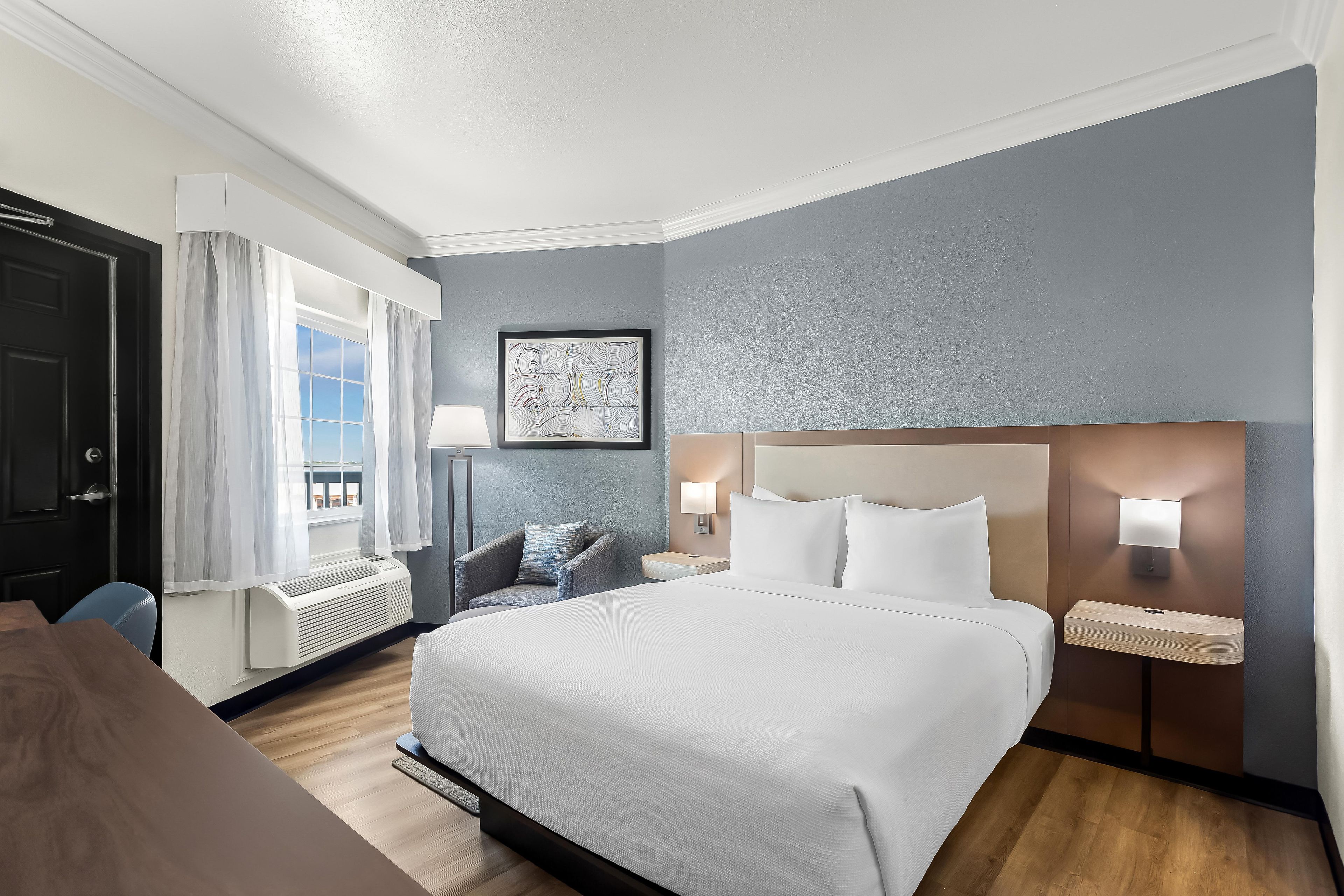A hotel room with a large bed, a desk, a chair, a window with curtains, a wall-mounted lamp, and a framed picture.