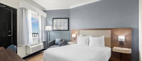A hotel room with a large bed, a desk, a chair, a window with curtains, a wall-mounted lamp, and a framed picture.