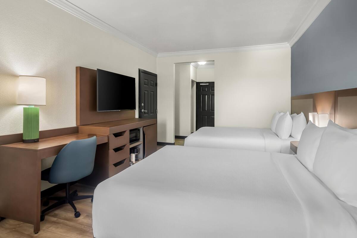 Quarto de hotel com cama, escrivaninha, cadeira, televisão e porta.