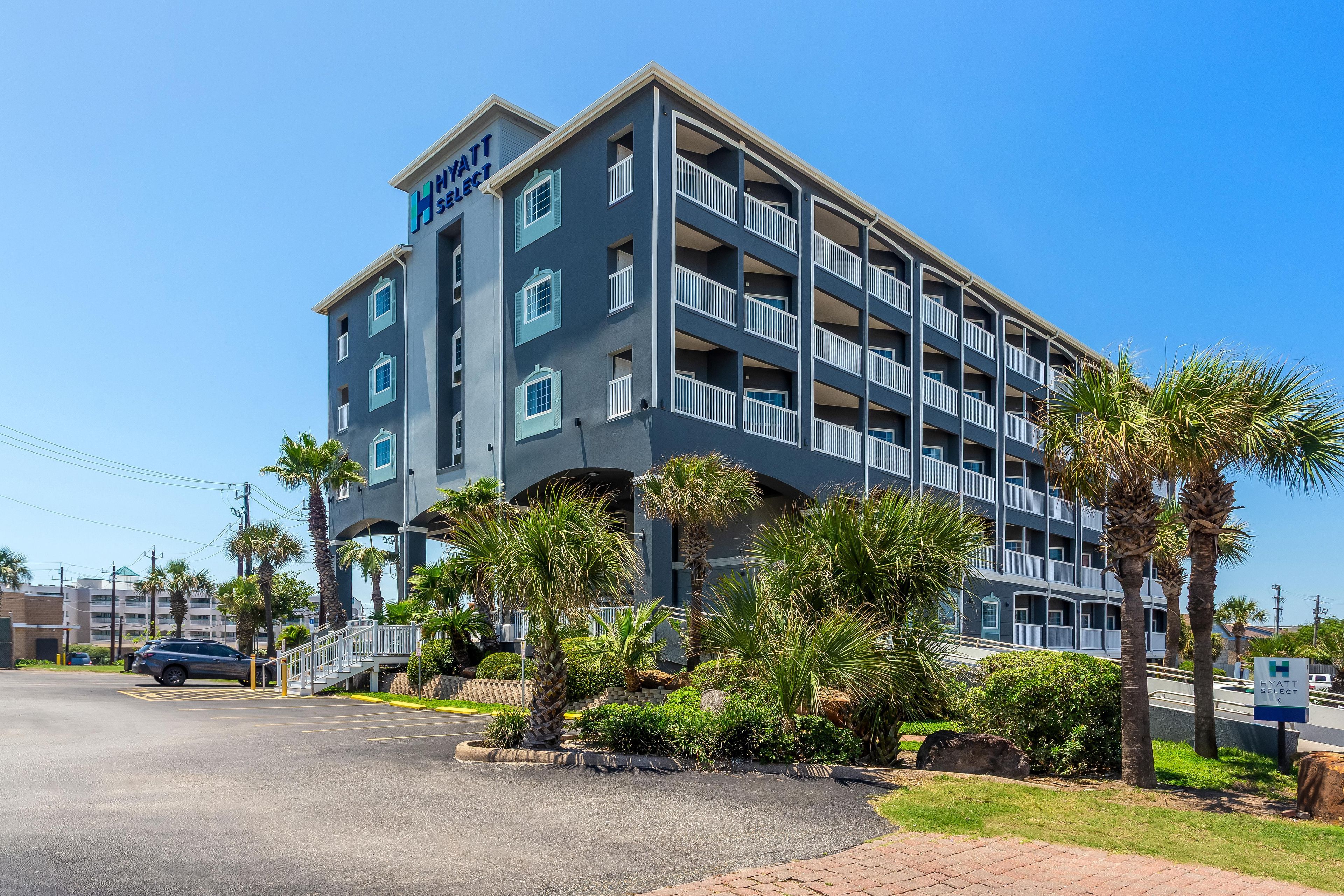 A multi-story hotel with a flat facade, balconies, and a parking area with palm trees.