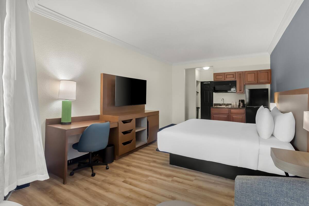 Quarto de hotel com cama, escrivaninha, cadeira, televisão e cozinha compacta.