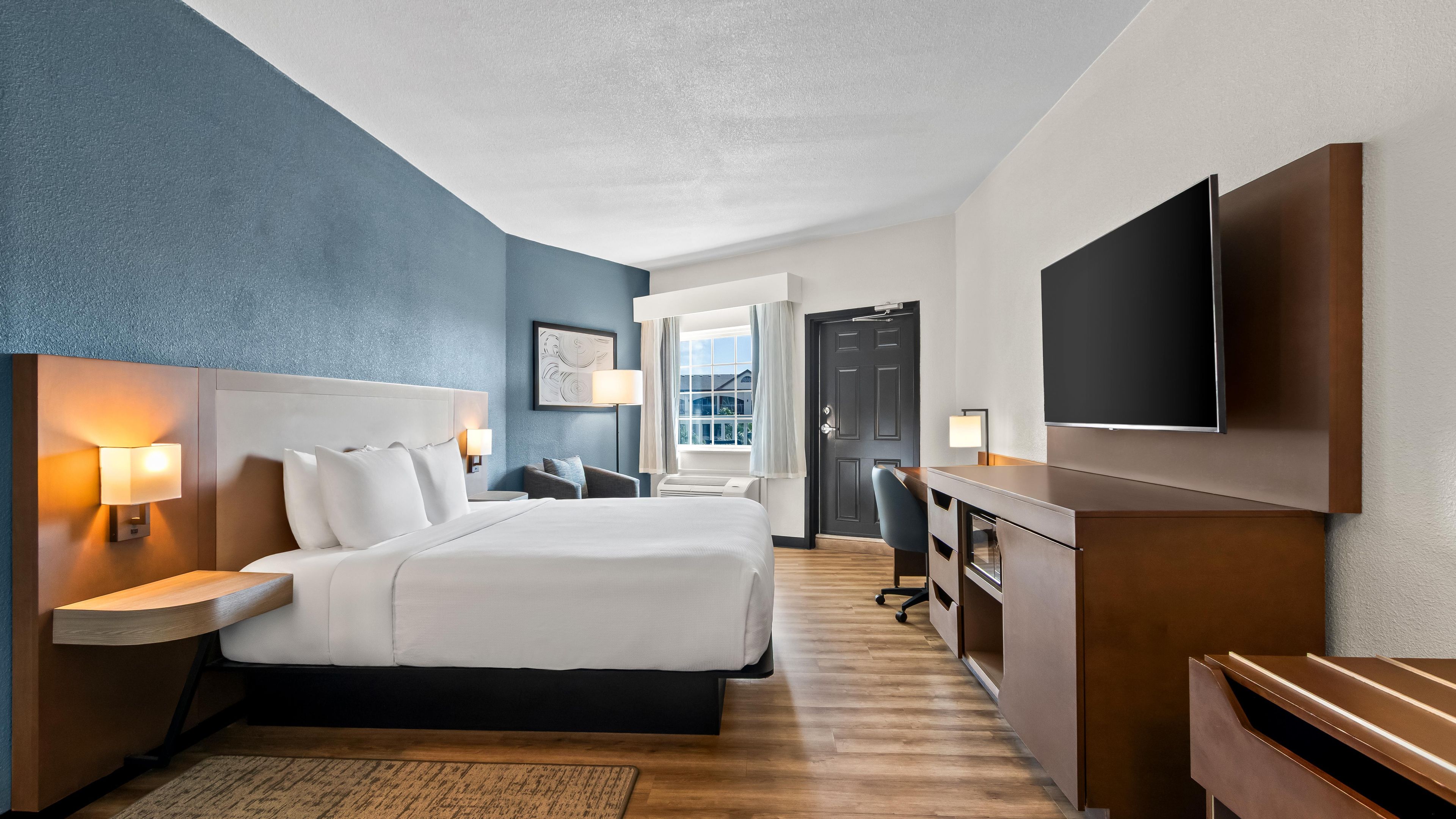 A hotel room with a large bed, bedside tables with lamps, a desk with a flat-screen TV, and a view of the outdoors through a window.
