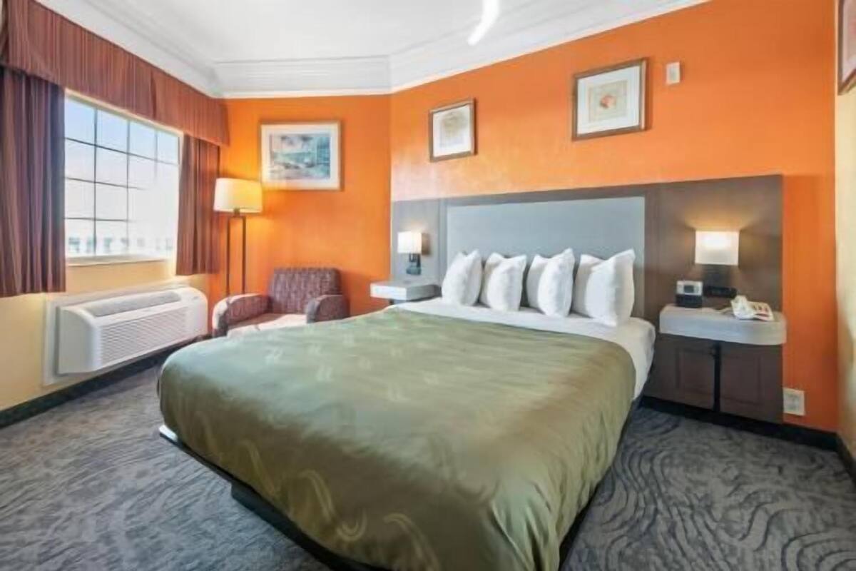 Quarto de hotel com uma cama grande, uma cadeira, uma escrivaninha e uma janela.