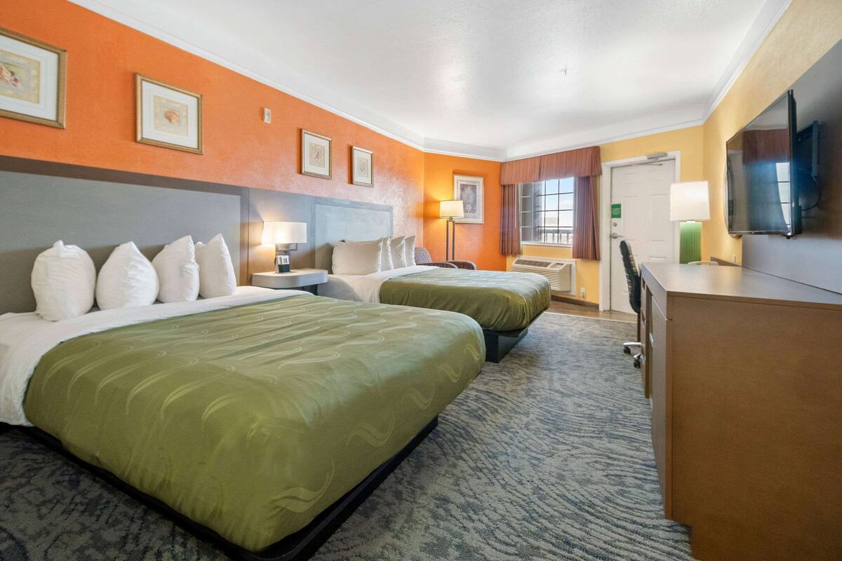 Quarto de hotel com duas camas, uma escrivaninha, televisão e janela com cortinas.