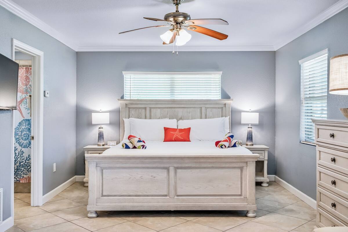 Quarto com cama branca, duas luminárias de apoio, ventilador de teto e janela com persianas.