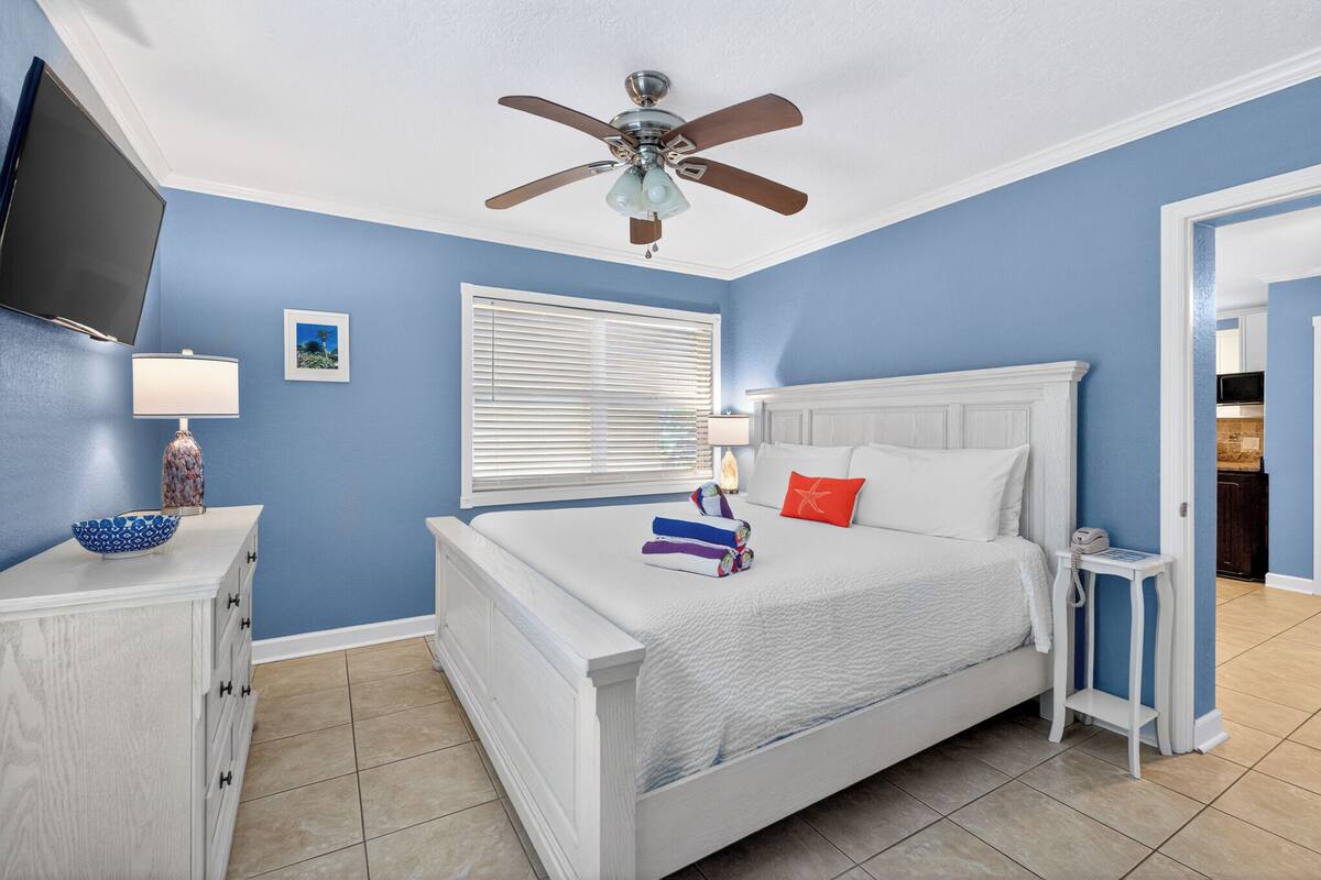 Quarto com cama, ventilador de teto, TV, janela com persianas, mesa de cabeceira e criado-mudo.