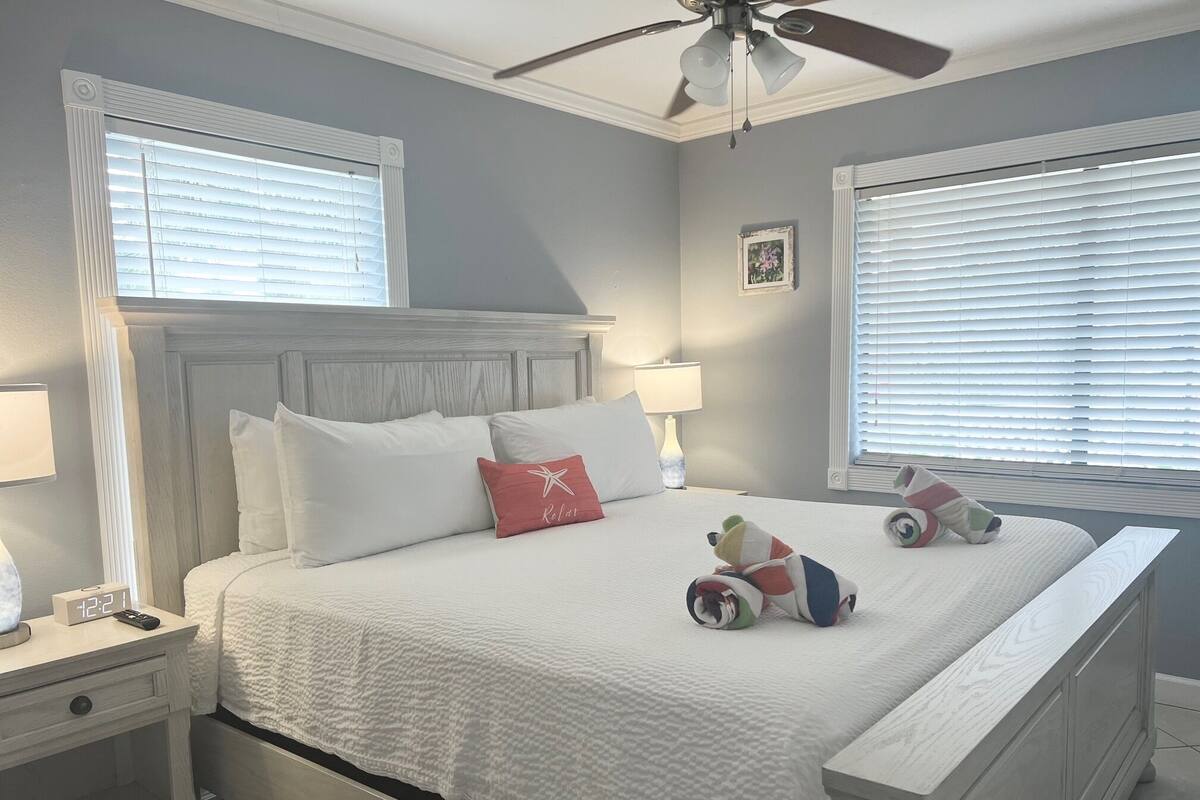 Quarto bem arrumado com cama, mesas de cabeceira, ventilador de teto e duas janelas com persianas.