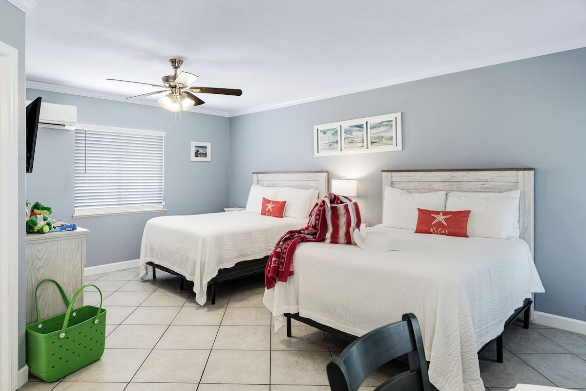Quarto com duas camas, um ventilador de teto, janela com persianas e um cesto verde.