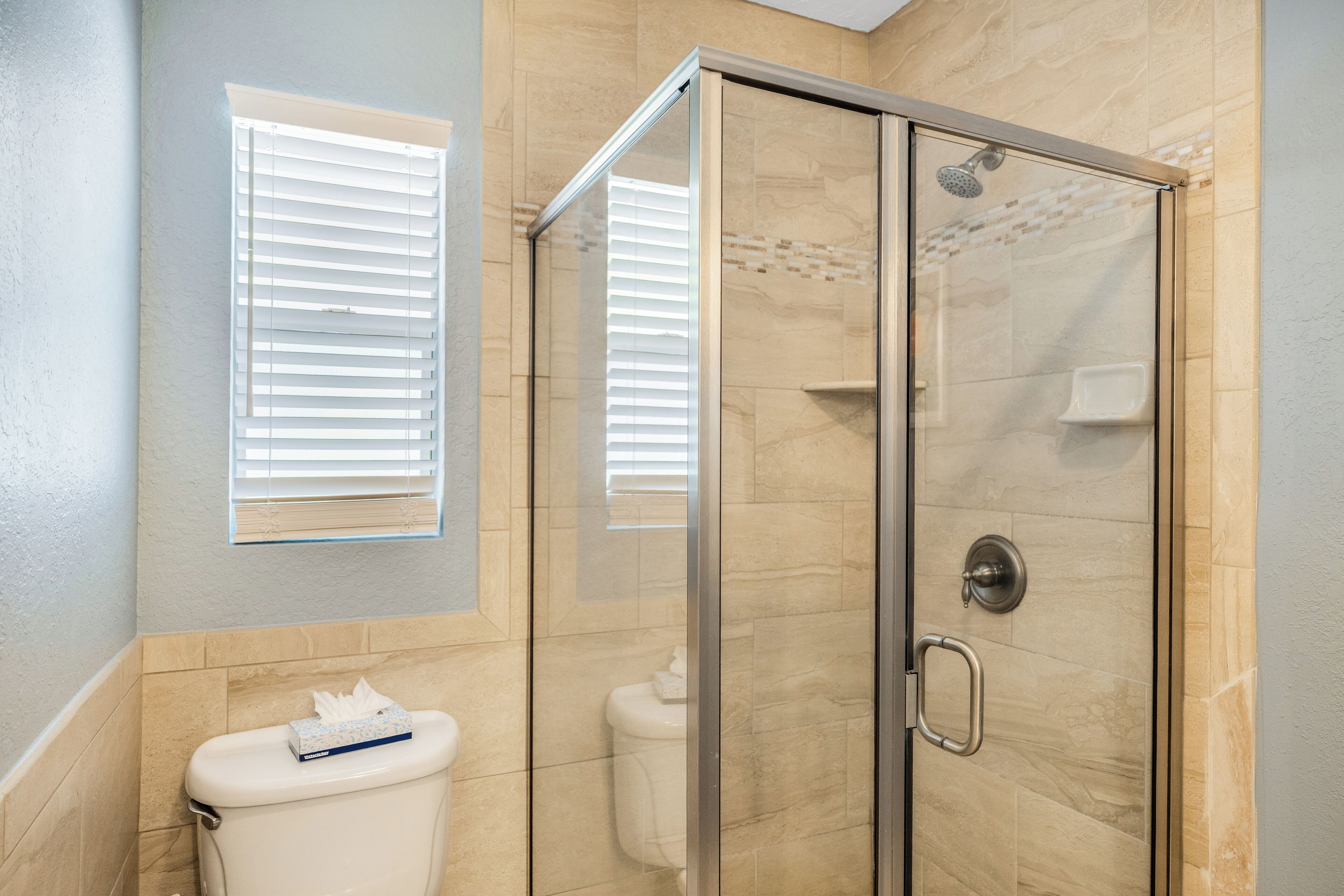 A bathroom with a glass-enclosed shower, a toilet, and a window with blinds.