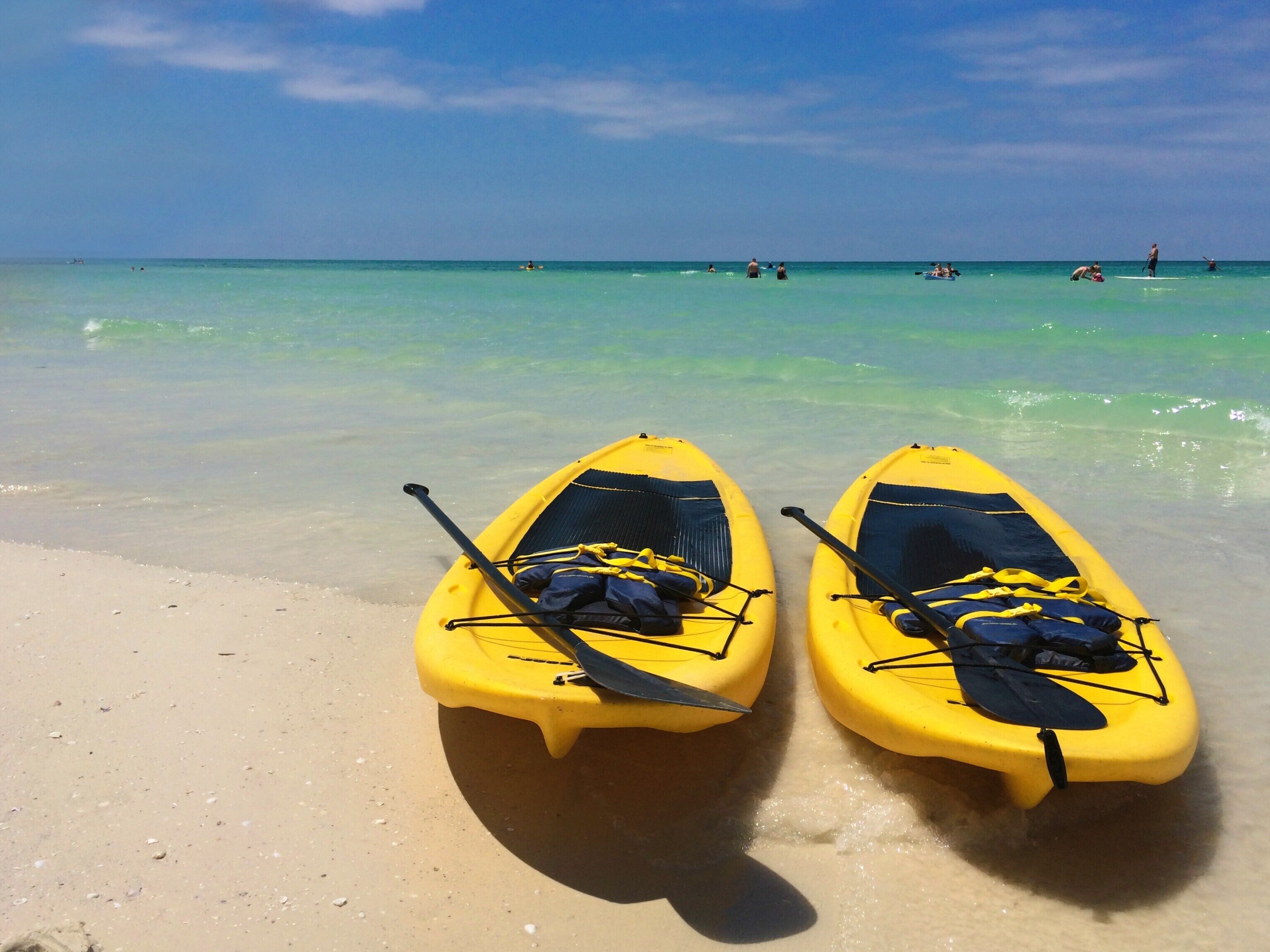 Dos kayaks amarillos con remos y chalecos salvavidas en una playa arenosa con agua turquesa cristalina y algunas personas a lo lejos.