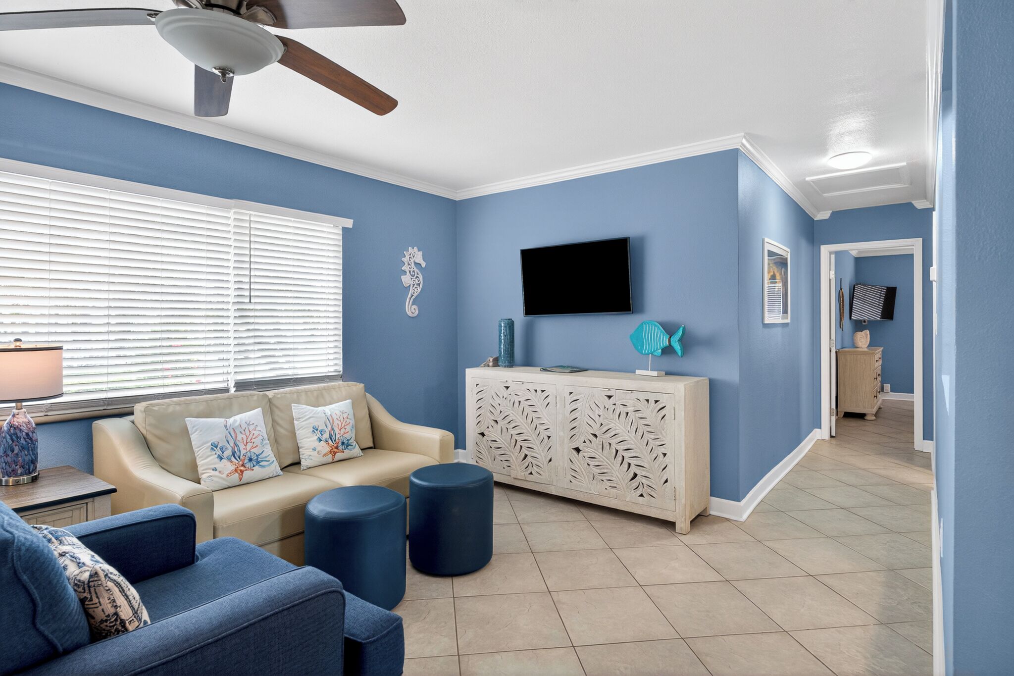 Una sala de estar con un sofá color beige, sillones azules, un aparador blanco y un ventilador de techo.