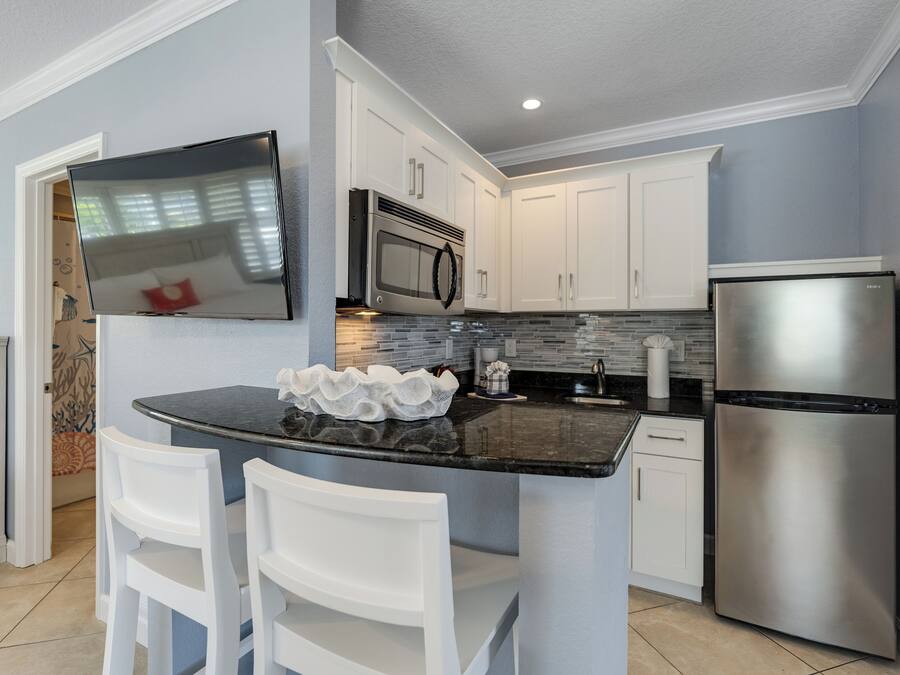 A modern kitchen with white cabinets, a stainless steel refrigerator, a microwave, and a flat-screen TV mounted above the island.