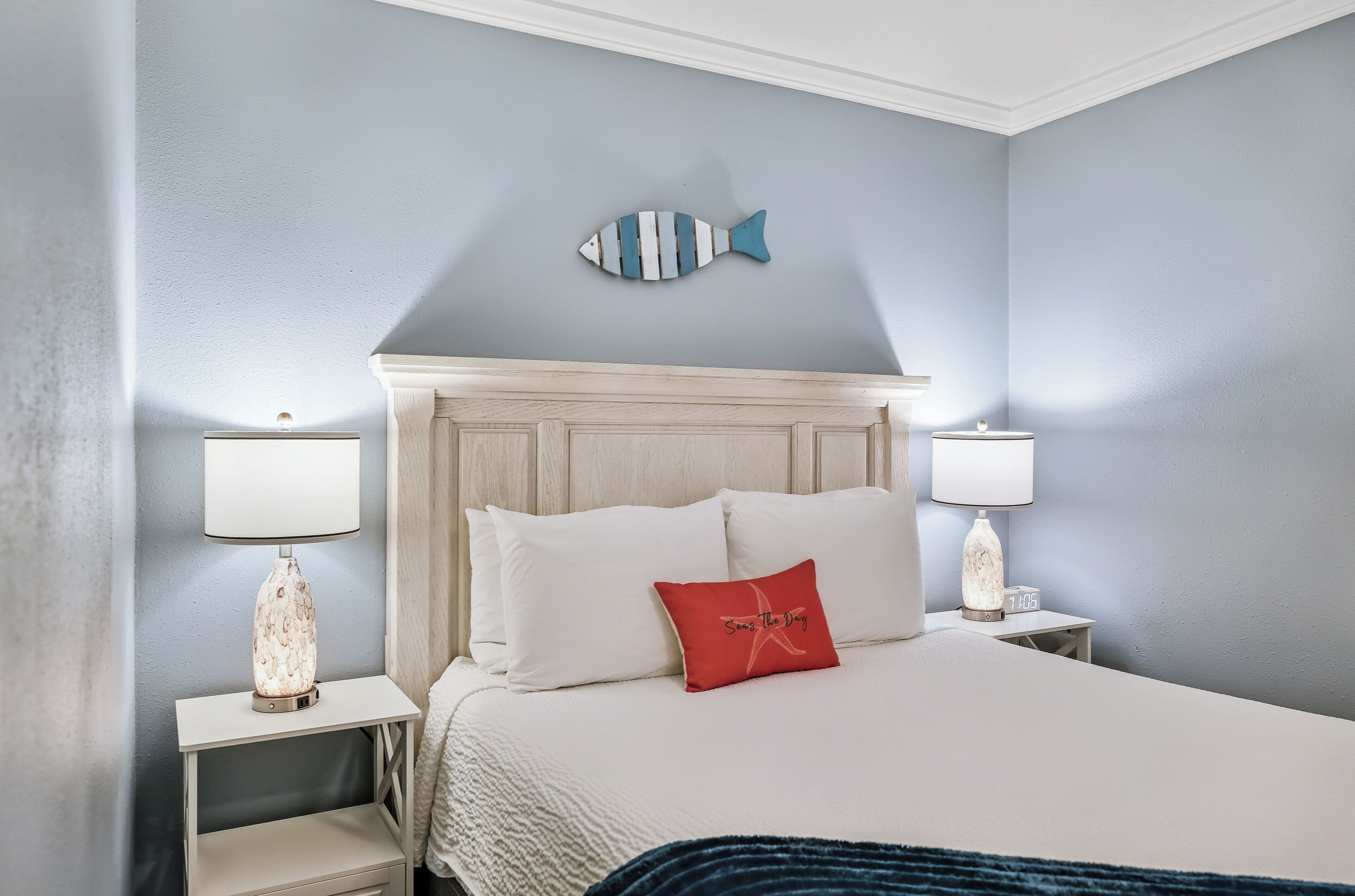 Un lit bien fait, avec du linge de lit blanc, un coussin rouge et une décoration murale représentant un poisson bleu. Il y a deux tables de chevet avec des lampes, et une petite horloge sur l’une d’elles.