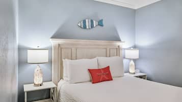 Un lit bien fait, avec du linge de lit blanc, un coussin rouge et une décoration murale représentant un poisson bleu. Il y a deux tables de chevet avec des lampes, et une petite horloge sur l’une d’elles.