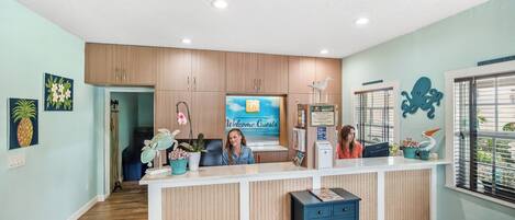 Un espace d’accueil avec un comptoir en bois, une pancarte indiquant « Bienvenue à nos invités » et des éléments décoratifs, comme un tableau représentant un ananas et une sculpture d’oiseau.