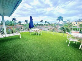 A patio with a table and chairs, surrounded by a white metal fence, and palm trees in the background.