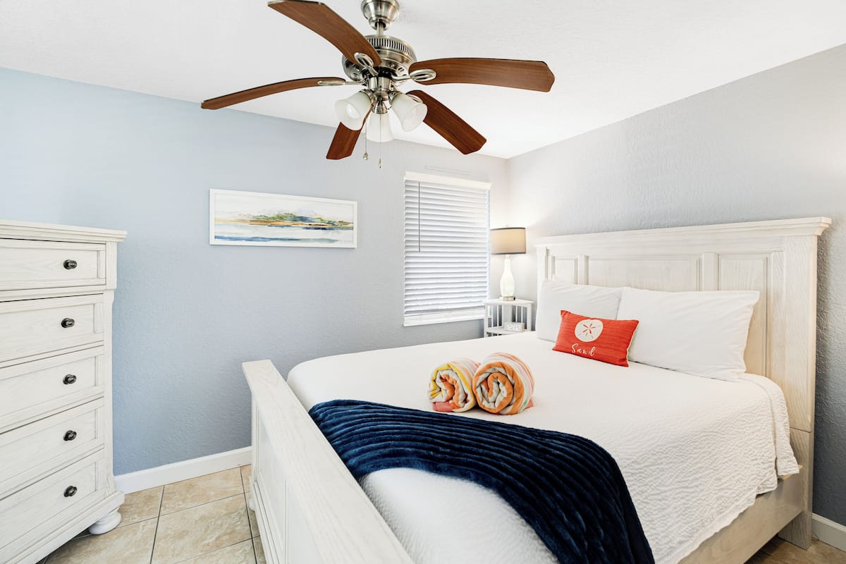 Quarto com cama, ventoinha no teto, cômoda, janela com persianas e um quadro na parede.