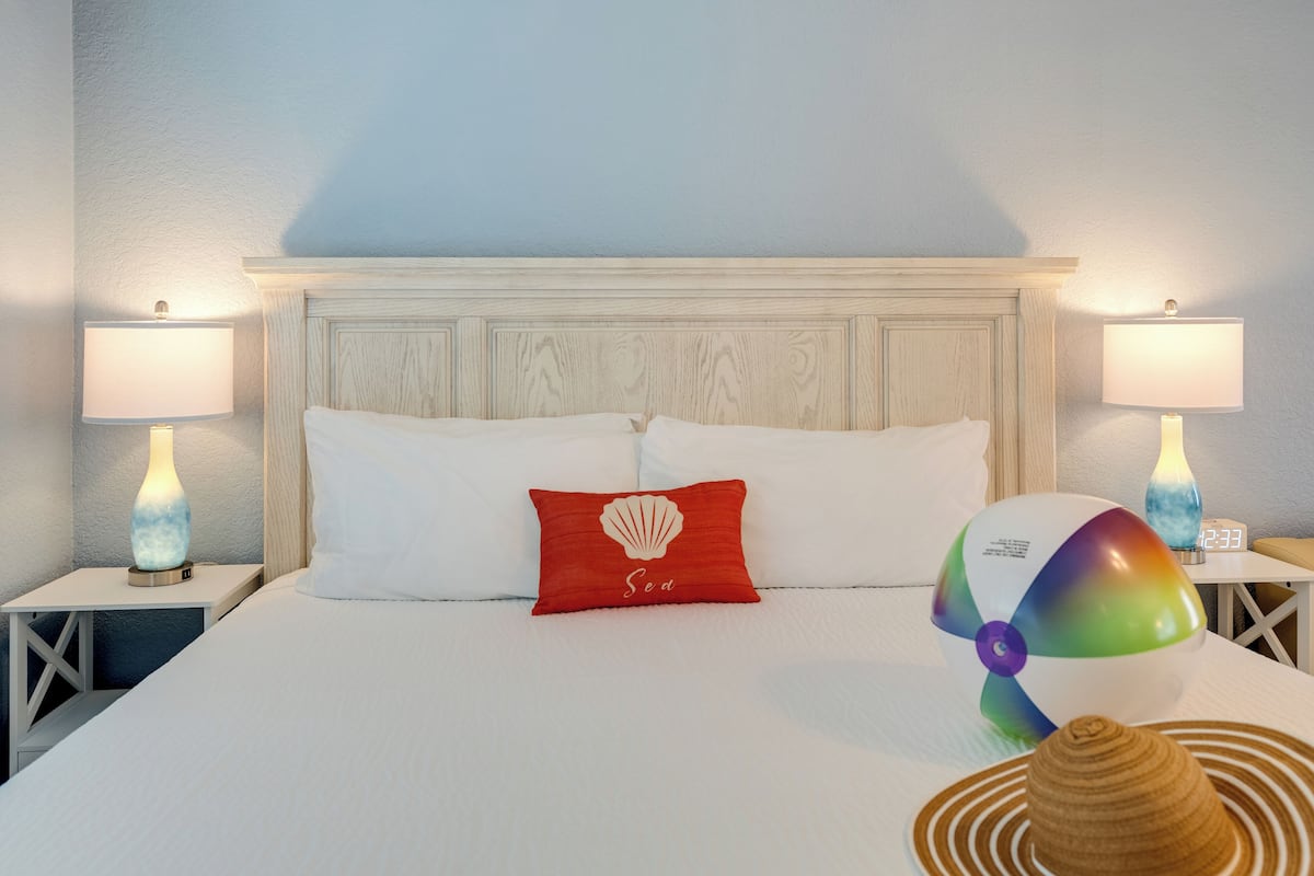 Cama bem arrumada com lençóis brancos, um travesseiro vermelho com desenho de concha e uma boia de praia ao lado.
