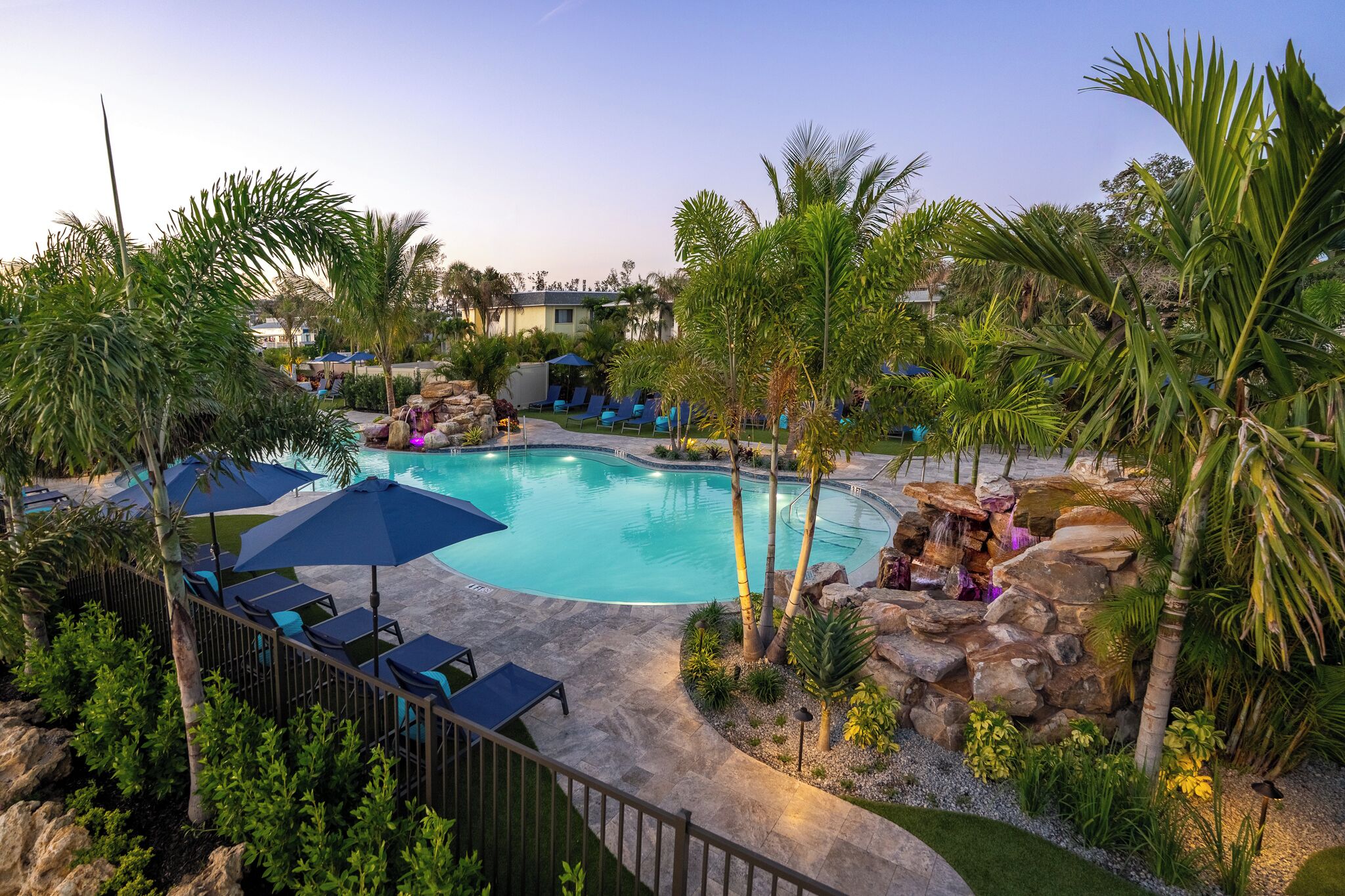 A pool area with lounge chairs and umbrellas, surrounded by palm trees and a waterfall feature.
