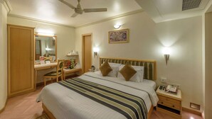 A hotel room with a bed, a desk, a chair, a mirror, a bedside table with a phone, and a ceiling fan. - Comfort Inn President (Ahmedabad)