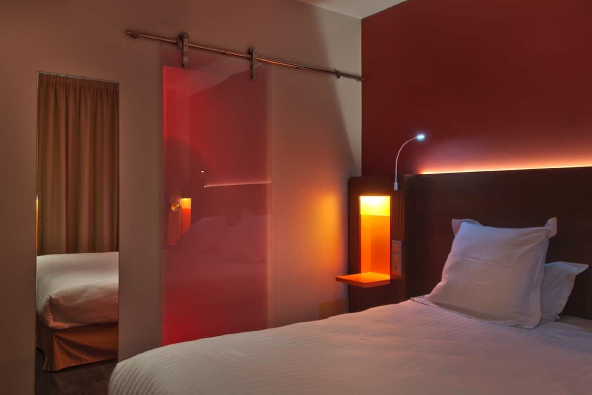 A hotel room with a bed, a nightstand, a wall-mounted lamp, and a curtain.
