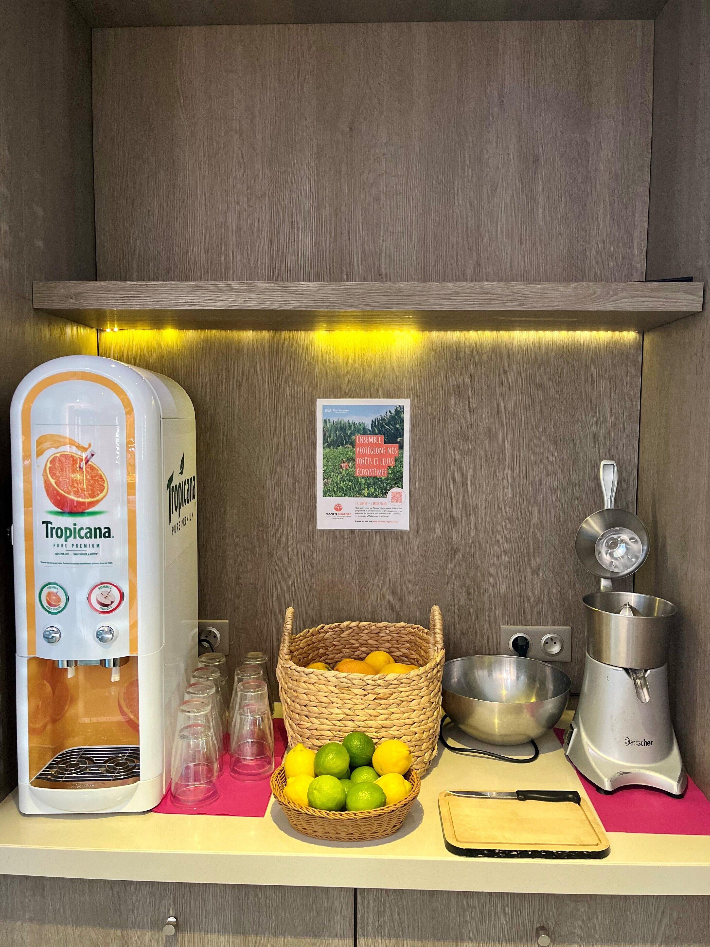 A kitchen counter with a water dispenser, a basket of citrus fruits, a mixing bowl, a juicer, and a cutting board.