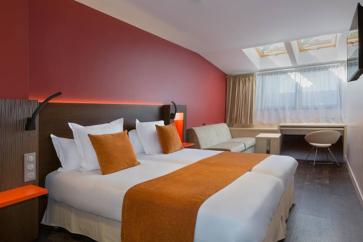 A hotel room with a large bed, a desk, a chair, and a skylight.