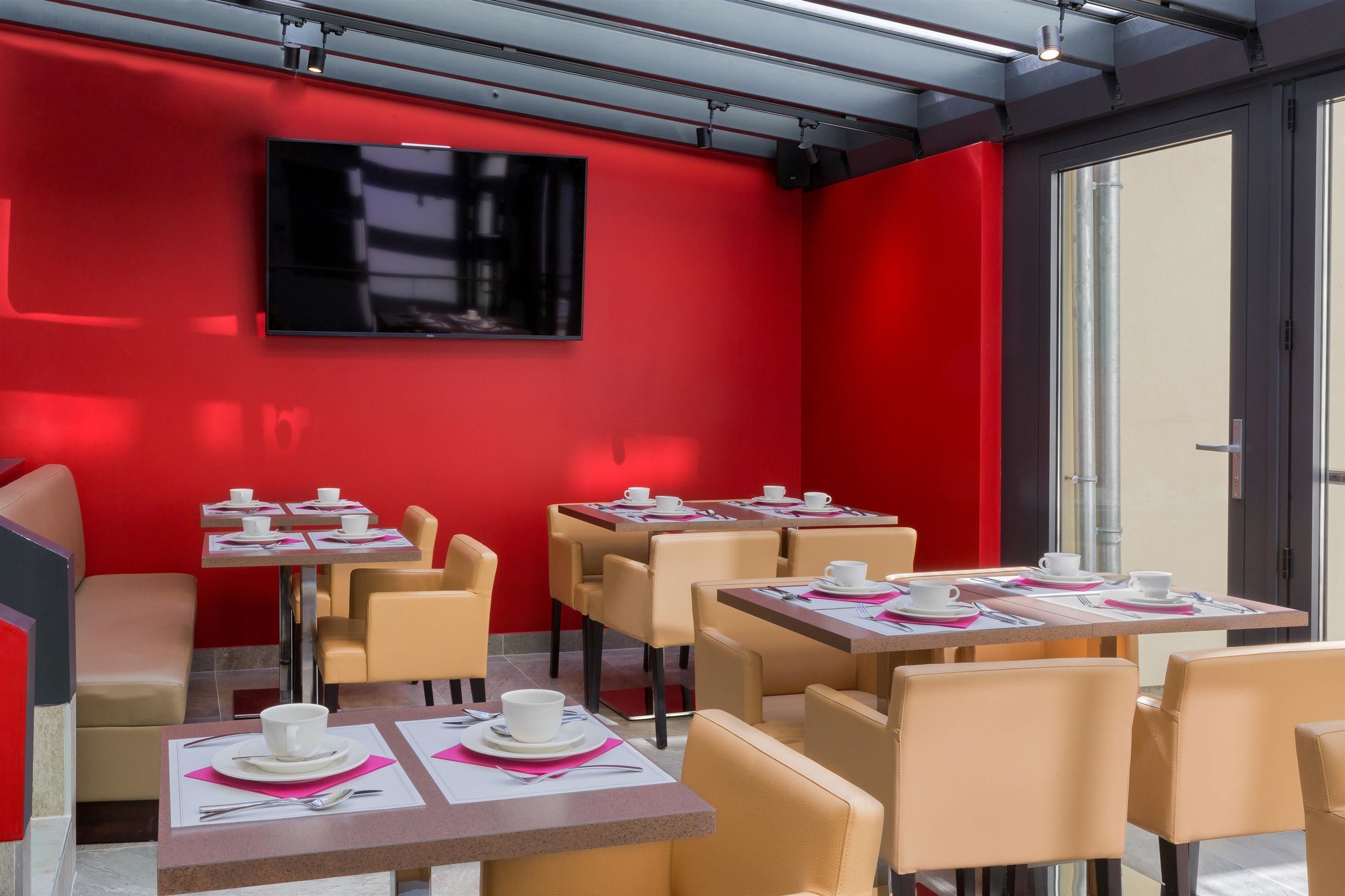 Una cafetería moderna con paredes rojas, varias mesas con tazas y platillos blancos, y un televisor de pantalla plana montado en la pared.