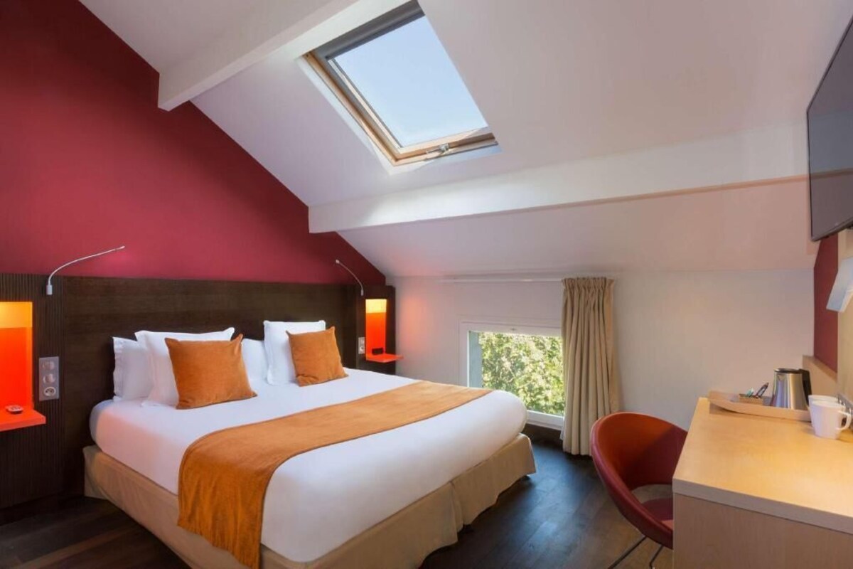 A hotel room with a bed, a desk, a chair, a desk lamp, a nightstand, a window, and a skylight.