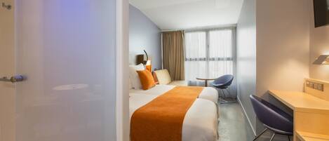 Una habitación de hotel con cama, escritorio, silla, televisor y ventana con cortinas.