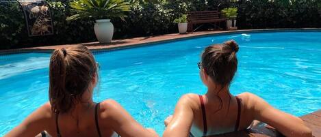 Two people relaxing in a swimming pool with a tiled deck, surrounded by lush greenery and a large planter.