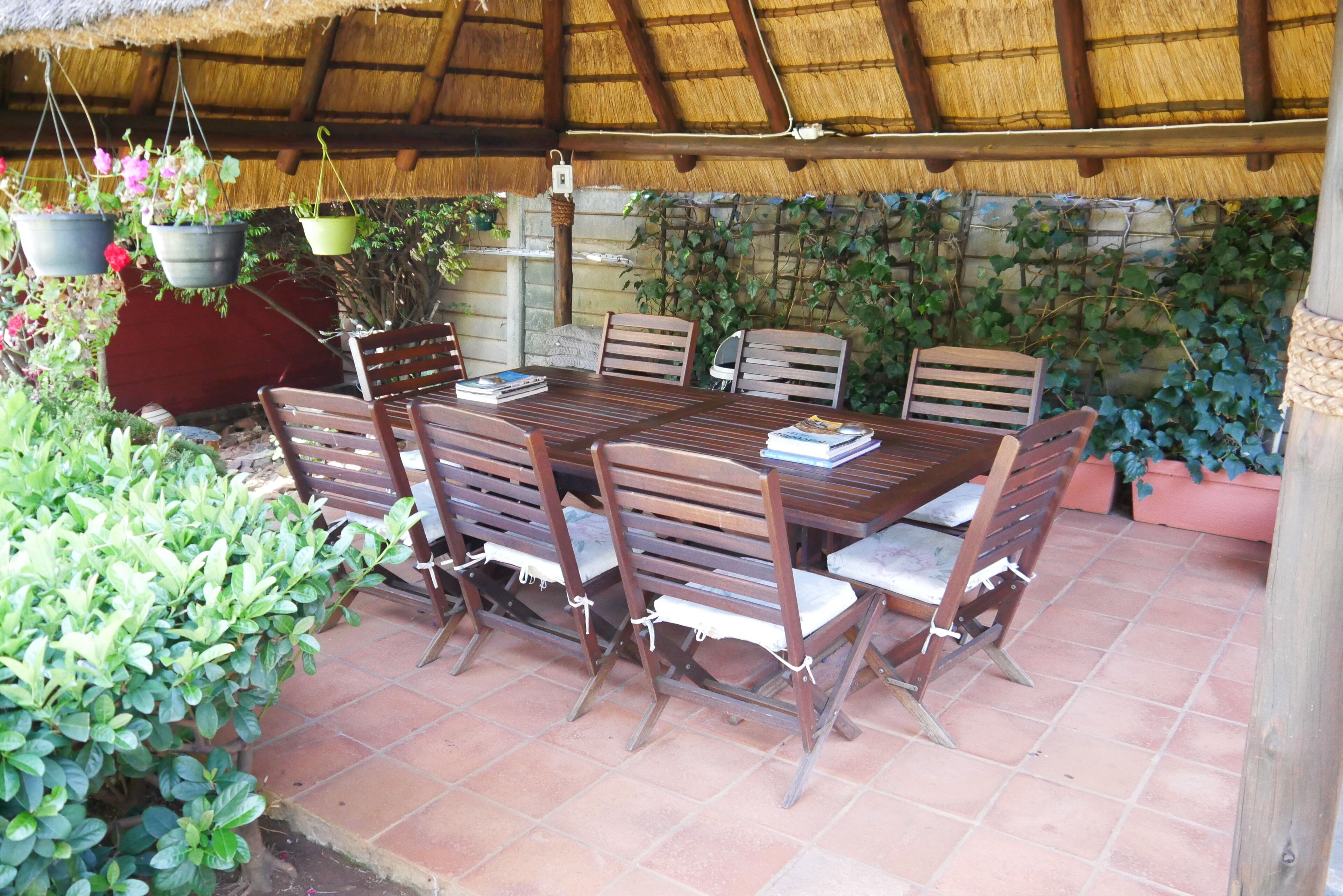 An outdoor patio with a wooden table and chairs, surrounded by potted plants and a thatched roof.