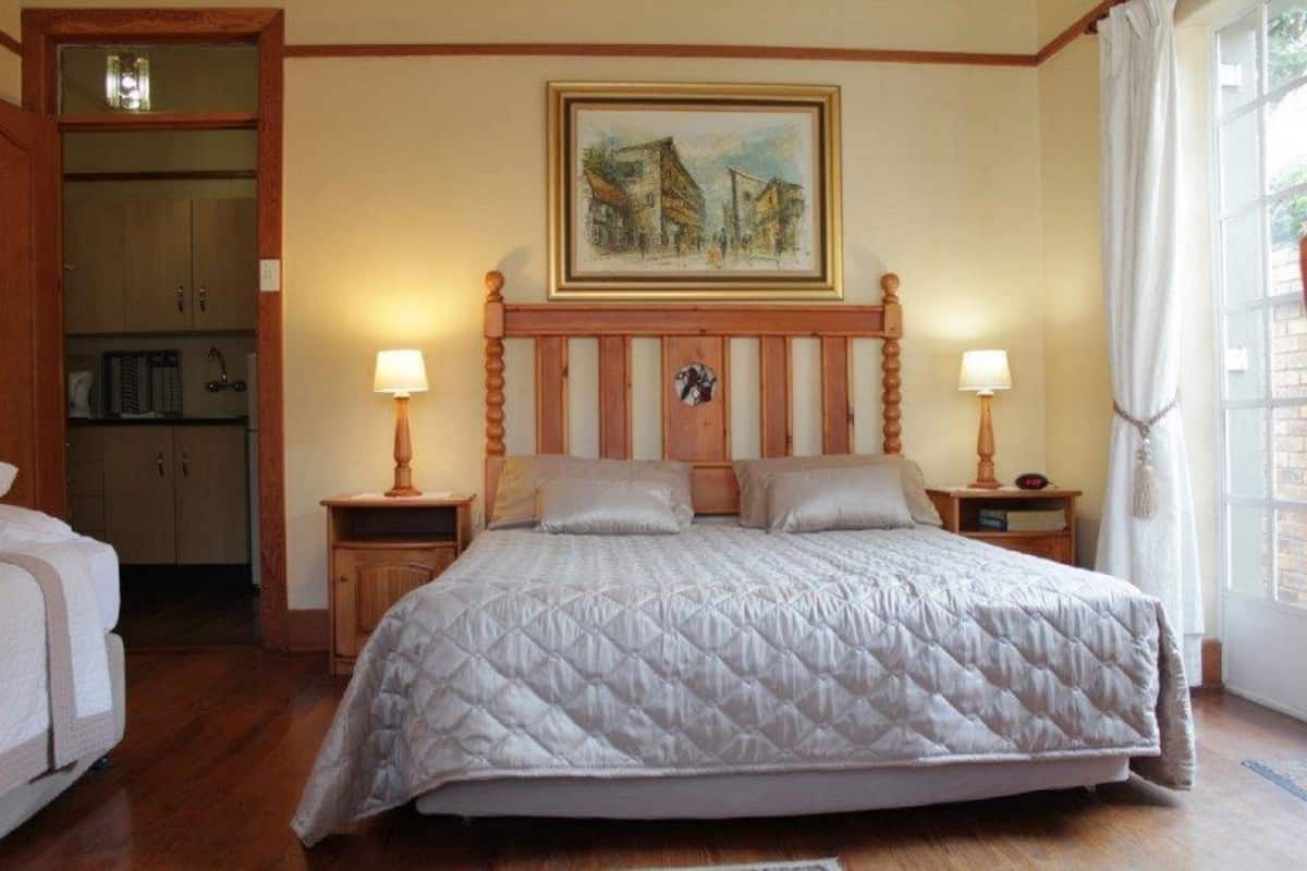 Un dormitorio con cama, cabecera de madera, dos mesitas de noche con lámparas, un cuadro en la pared y una ventana con cortinas.