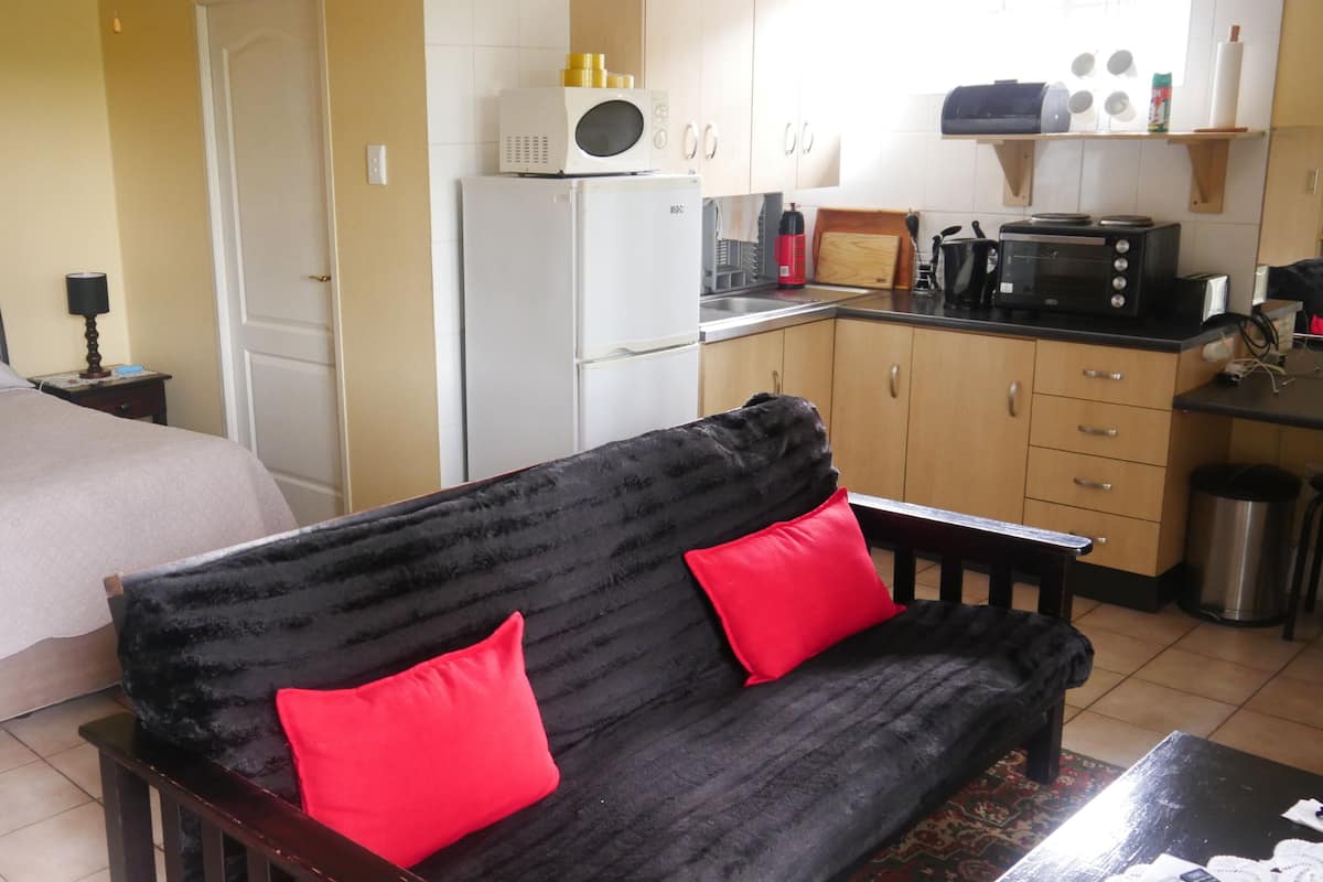 Una habitación de hotel compacta con cama, sofá con cojines rojos, kitchenette y pequeña mesa de comedor.