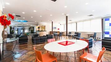 Eine geräumige Lobby mit einem runden weißen Tisch, orangefarbenen Stühlen, schwarzen Sofas und einem großen Fenster, das für natürliches Licht sorgt.