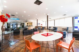 A spacious lobby with a round white table, orange chairs, black sofas, and a large window providing natural light.