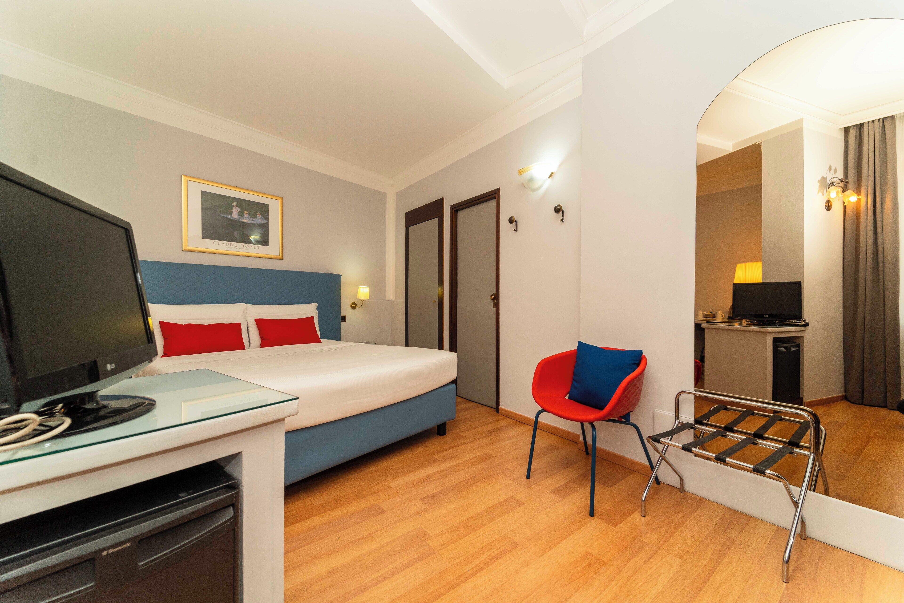 A hotel room with a bed, a desk with a television, a chair, a mirror, and a small kitchenette.