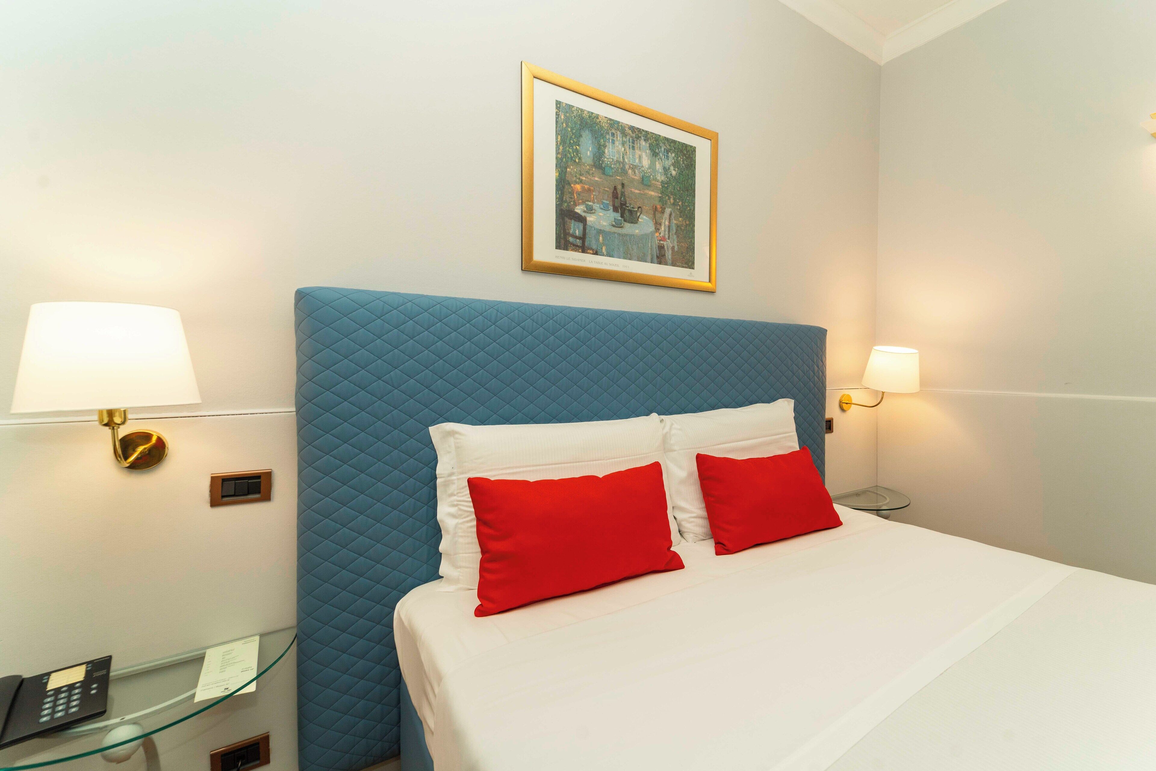 Une chambre d’hôtel avec un lit, deux oreillers rouges, une tête de lit bleue et un tableau encadré au mur.