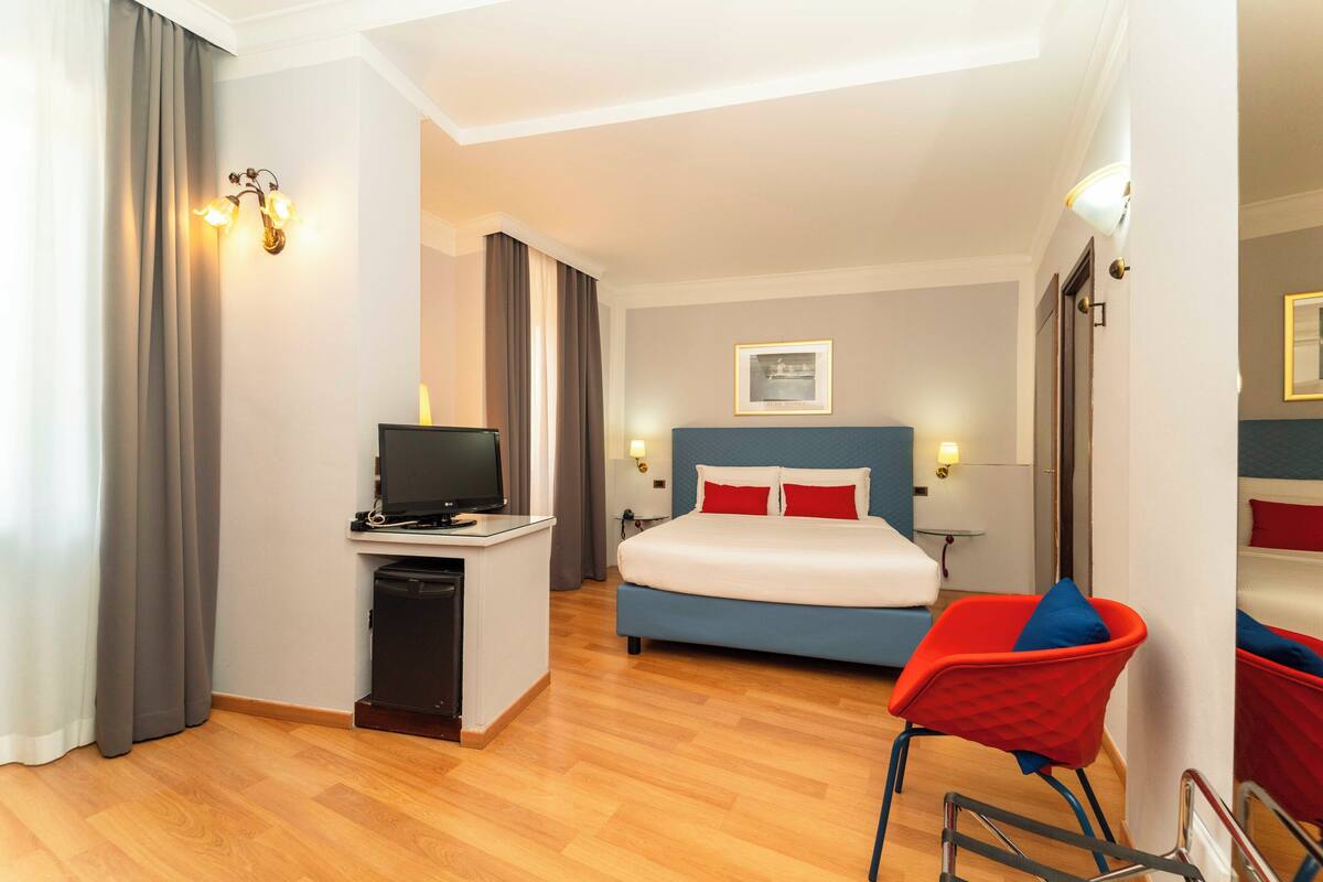 A hotel room with a bed, a desk with a computer, a chair, and a television.