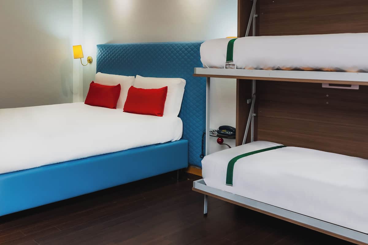 A hotel room with a blue headboard, a single bed with white bedding and red pillows, and a bunk bed with white bedding.