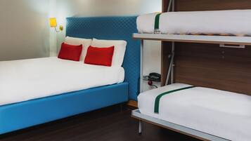 Habitación de hotel con cabecera azul, cama individual con ropa blanca y almohadas rojas, y literas con ropa blanca.