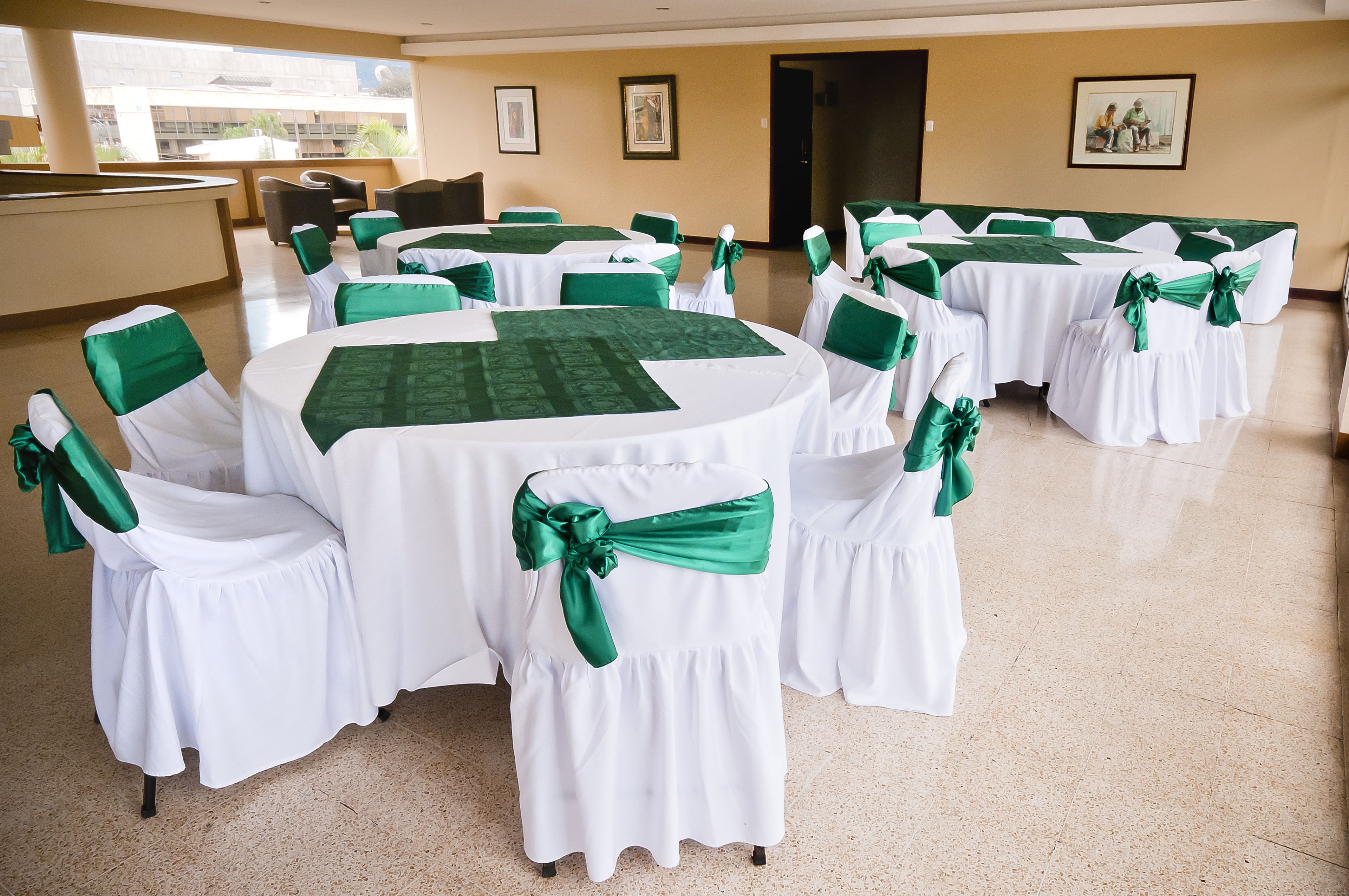 Un salón de conferencias con mesas redondas cubiertas con manteles blancos y cintas verdes, dispuestas en forma de U.
